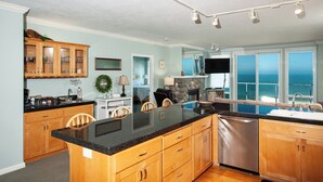 A modern kitchen with wooden cabinets, a black countertop, and a dishwasher. There is a dining area with chairs and a table, and a large window providing a view of the ocean. - Promo! Stunning Oceanfront Condo, Wifi, HDTV, Hot Tub, Indoor Pool and More! (Depoe Bay)
