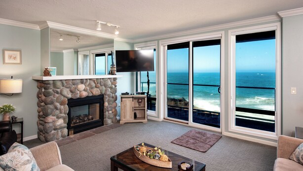 A living room with a stone fireplace, a flat-screen TV, and large windows overlooking the ocean. - Promo! Stunning Oceanfront Condo, Wifi, HDTV, Hot Tub, Indoor Pool and More! (Depoe Bay)