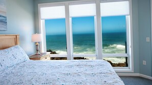 A bedroom with a large window overlooking the ocean, a bed with a floral bedspread, and a nightstand with a lamp. - Promo! Stunning Oceanfront Condo, Wifi, HDTV, Hot Tub, Indoor Pool and More! (Depoe Bay)