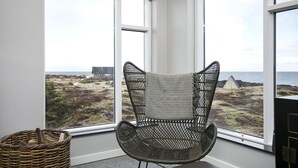 A corner room with a wicker chair facing a large window overlooking a coastal landscape.