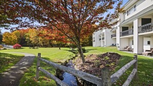A residential building with a grassy lawn, a small stream, and a wooden fence. - Ski-in/Ski-out Lodge Condo at Toll House Lift (Stowe)