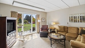 A living room with a TV, a sofa, a coffee table, and a chair. There is a glass door leading to a garden. - Ski-in/Ski-out Lodge Condo at Toll House Lift (Stowe)