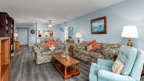 A living room with two floral-patterned sofas, a wooden coffee table, and a light blue armchair. There is a bookshelf, a lamp, and a ceiling fan. - Oceanfront view park on ocean nearby for kids (MYRTLE BEACH)