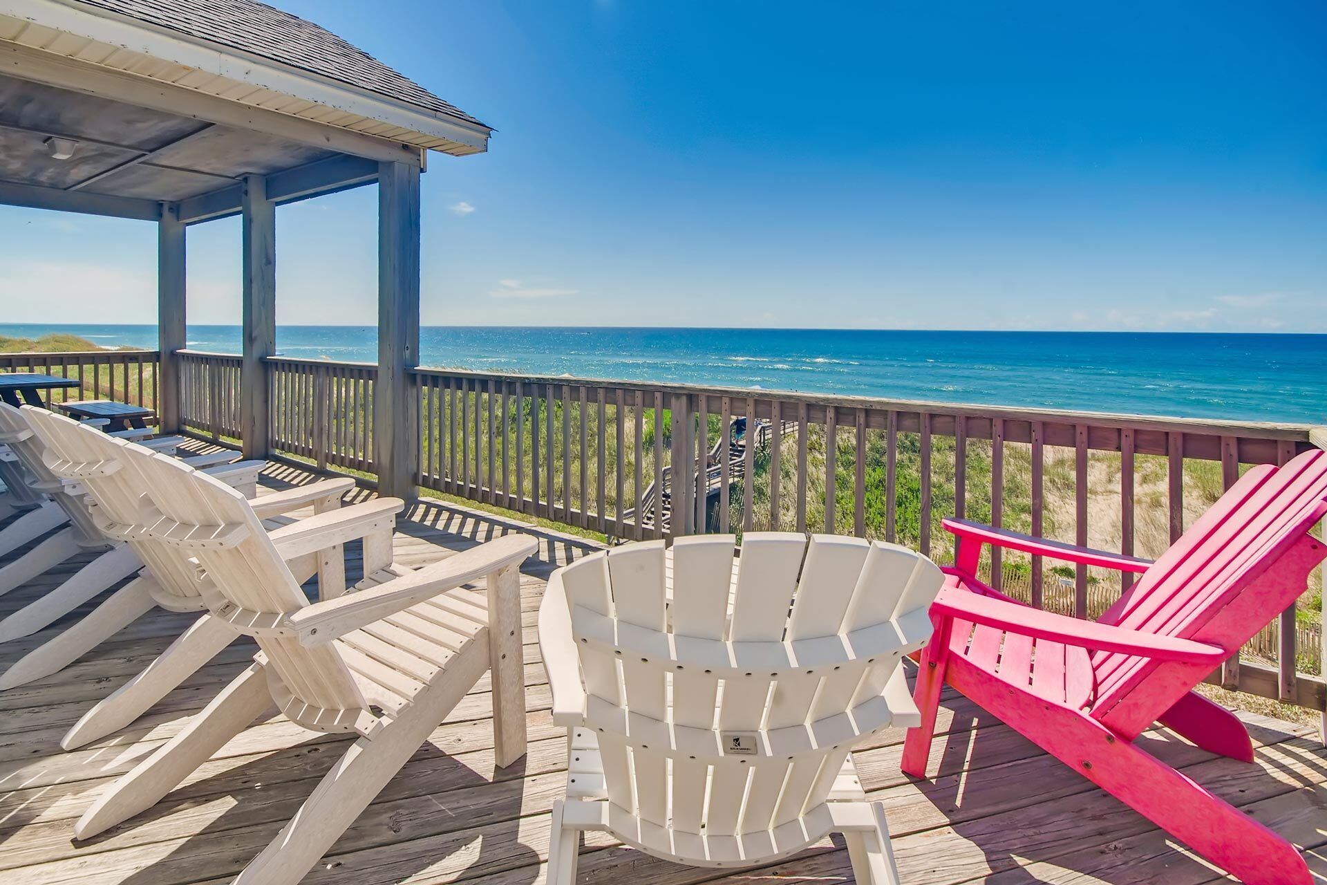 A wooden deck with white and pink Adirondack chairs overlooking the ocean.