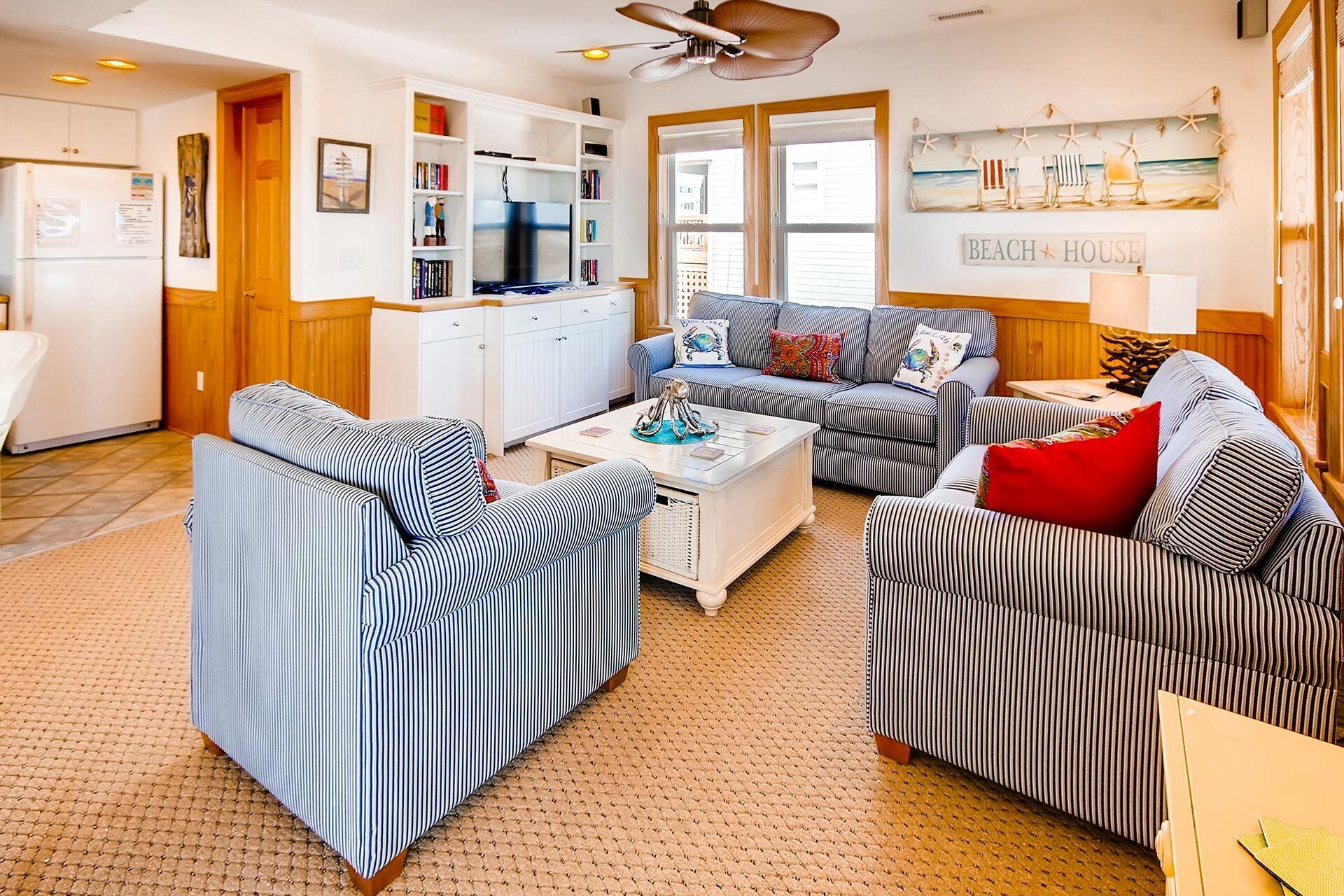 A living room with striped sofas, a coffee table, and a kitchen area with white cabinets and a refrigerator.