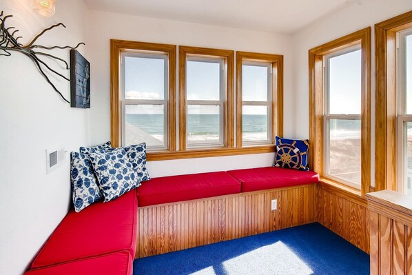 A corner seating area with a red bench and a blue rug, wooden window frames, and a view of the beach. - Oceanfront Dog Friendly Home in Hatteras w/ Pool, Hot Tub, Elevator, Game Room (Hatteras)