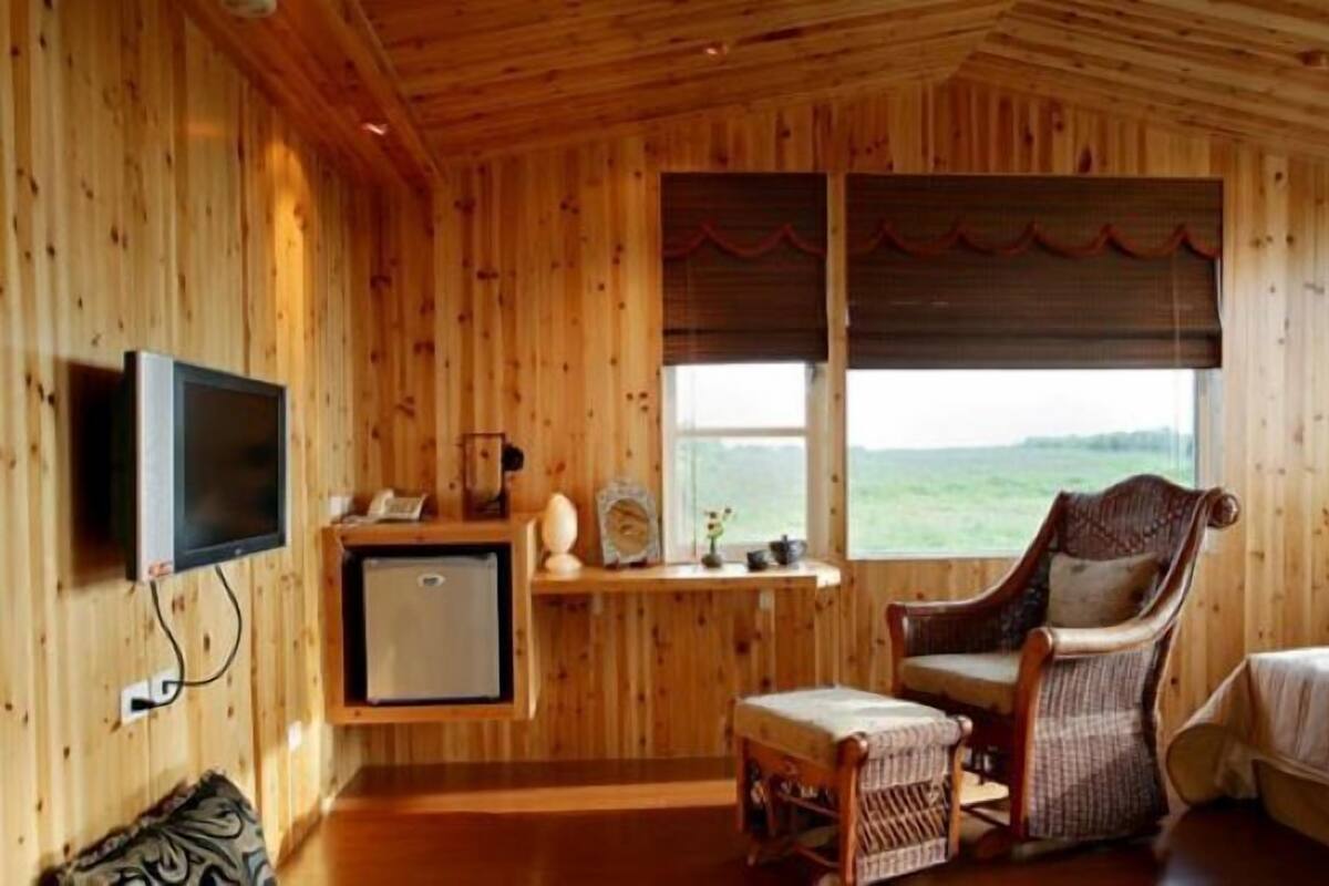 A wooden cabin room with a television, a small refrigerator, a chair, and a window with a view of the outdoors.
