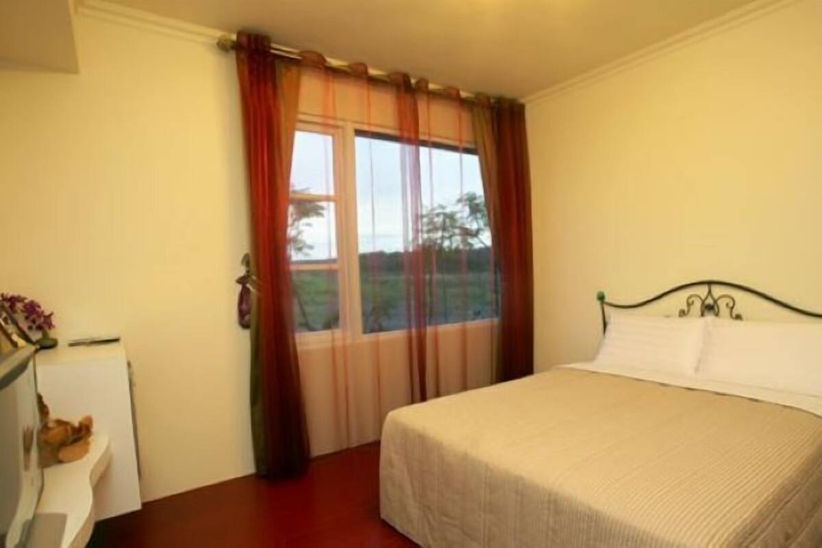 A bedroom with a bed, a window with curtains, a bedside table, and a small desk with a lamp.