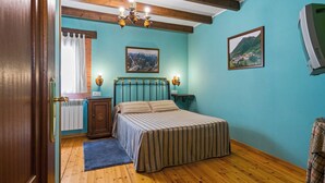 A bedroom with a wooden floor, a bed with striped bedding, a wooden headboard, a chandelier, two framed pictures on the wall, and a radiator.