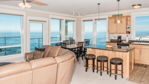 Una cucina moderna con vista sull'oceano, un'area pranzo con tavolo e sedie, e un angolo salotto con divano e sgabelli da bar.