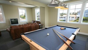 Een pooltafel met een witte knuppelbal, poolkeus en biljartballen. Op de achtergrond staat een bruine leren bank en een televisie.