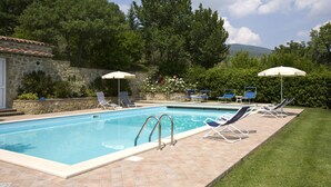 Ein Schwimmbecken mit Liegestühlen und Sonnenschirmen, umgeben von Grünflächen und einem Steinhaus.