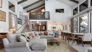 A spacious living room with a large sectional sofa, a dining area with wooden chairs, a kitchen in the background, and a staircase leading to an upper level. - Vail Village + Private Hot Tub, Game Room & Prestigious Forest Road Location (Vail)