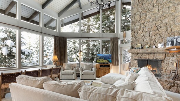 A spacious living room with a stone fireplace, large windows offering a view of snow-covered trees, and a cozy seating area with sofas and chairs. - Vail Village + Private Hot Tub, Game Room & Prestigious Forest Road Location (Vail)