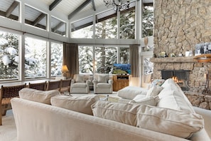 A spacious living room with a stone fireplace, large windows offering a view of snow-covered trees, and a cozy seating area with sofas and chairs.
