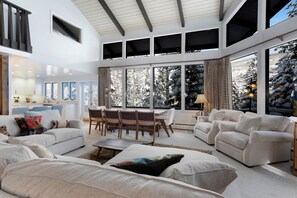 A spacious living room with a high ceiling, large windows, and a view of snow-covered trees.