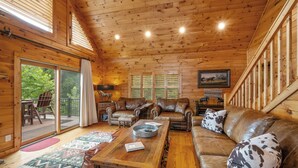 A cozy cabin interior with wooden walls, a leather sofa, a wooden coffee table, and a view of the outdoors through large windows. - Brook Haven: Private Cabin, Great Views, Hot Tub, WiFi, Fireplace (Ferguson)