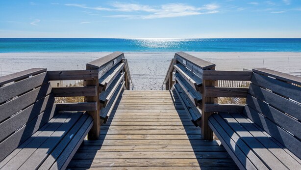 Ein hölzerner Steg führt zu einem Strand, der unter blauem Himmel liegt.
