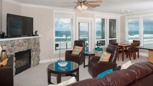 A living room with a stone fireplace, leather furniture, a flat-screen TV, and large windows overlooking the ocean. - Promo! Third Floor Corner Condo, Oceanfront, Private Hot Tub, Pool, Free Wifi! (Lincoln City)