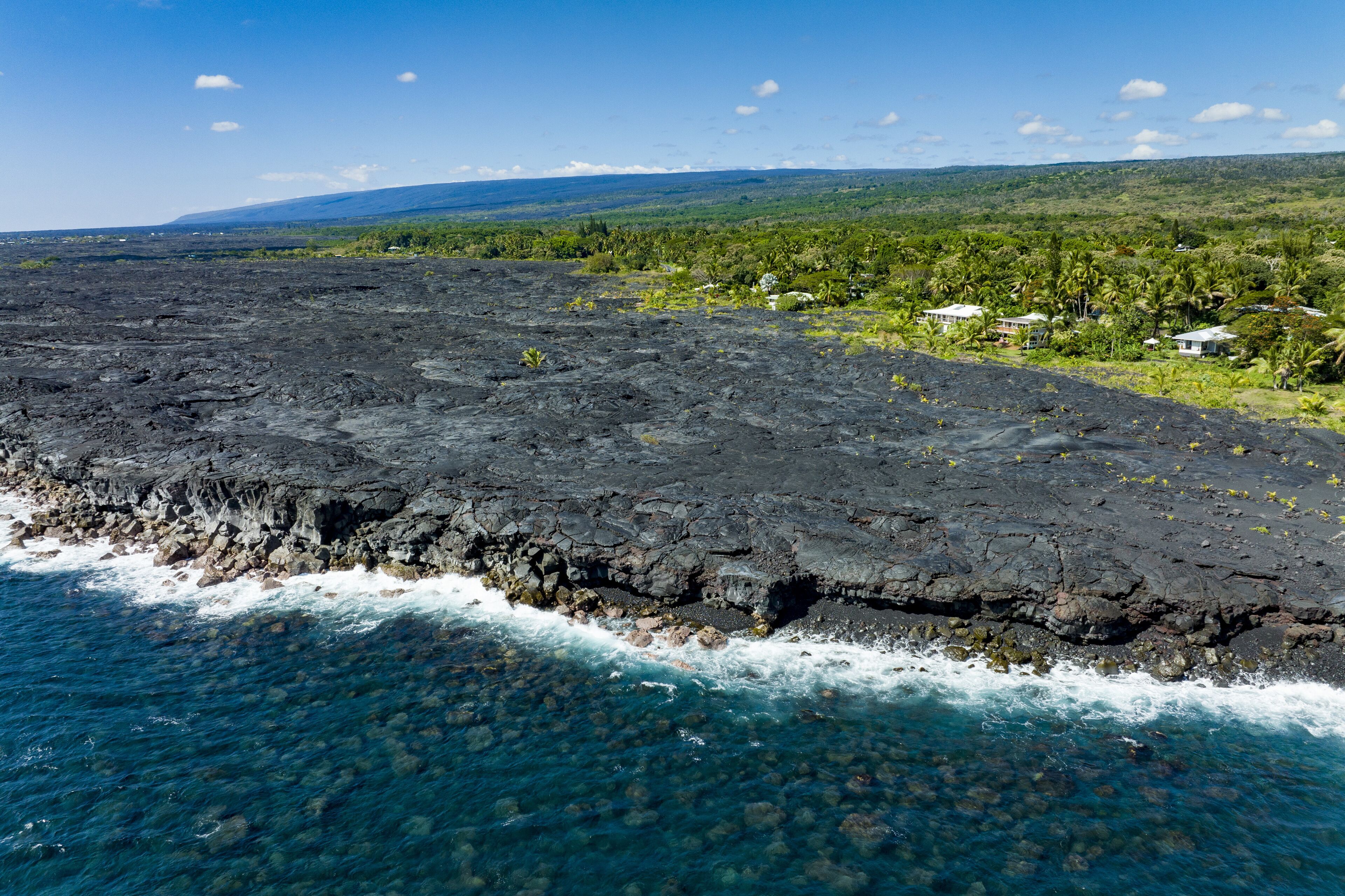 A coastal landscape with a rocky shoreline, clear blue waters, and a backdrop of lush greenery and distant hills.
