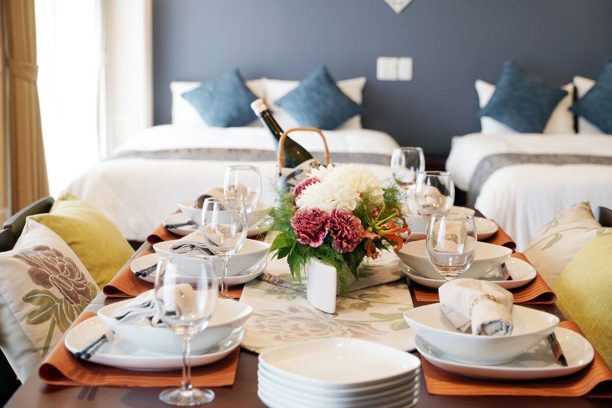 A neatly set dining table with plates, glasses, and a bottle of champagne, surrounded by pillows and a bed in the background.