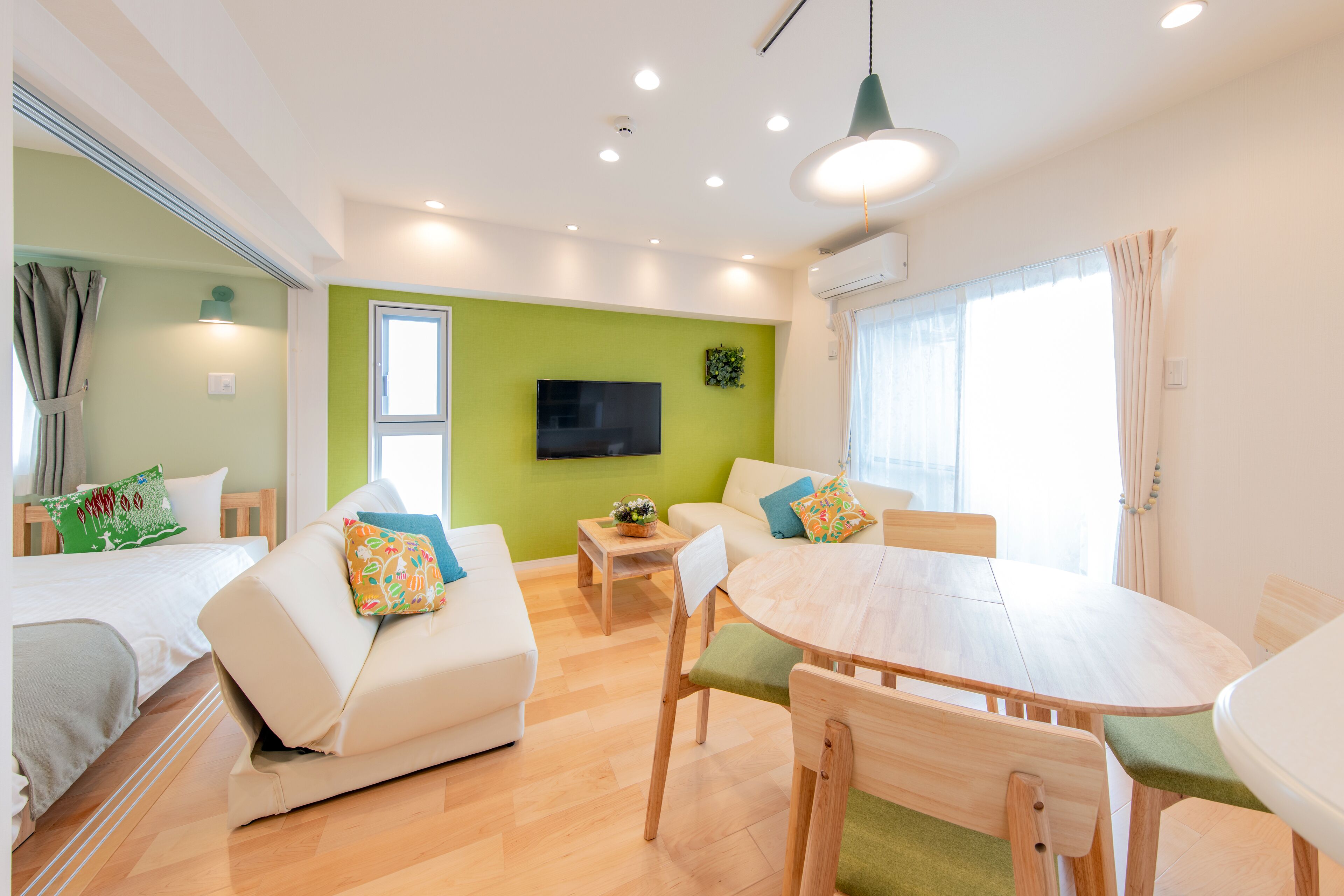 A modern, well-lit room with a bed, two white sofas with colorful cushions, a wooden dining table, and a TV mounted on the wall.