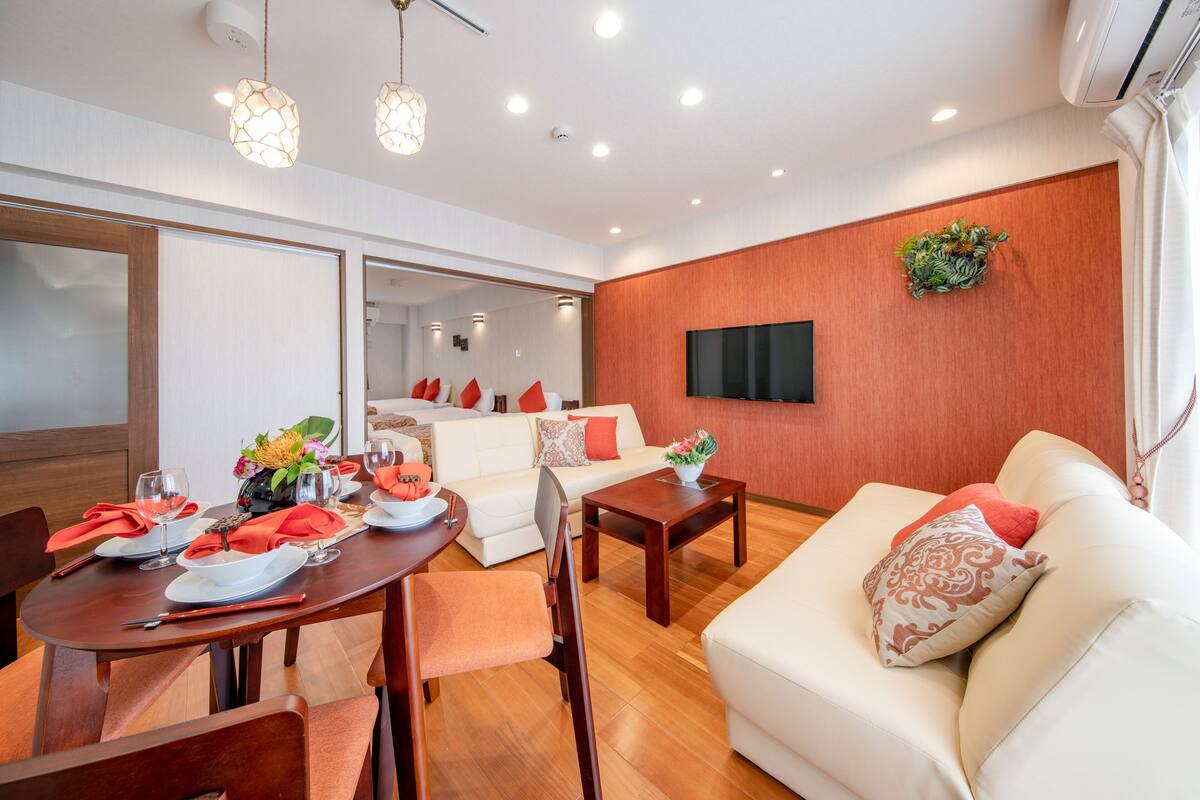 A modern living room with a dining area, white sofas, wooden flooring, and a flat-screen TV mounted on the wall.