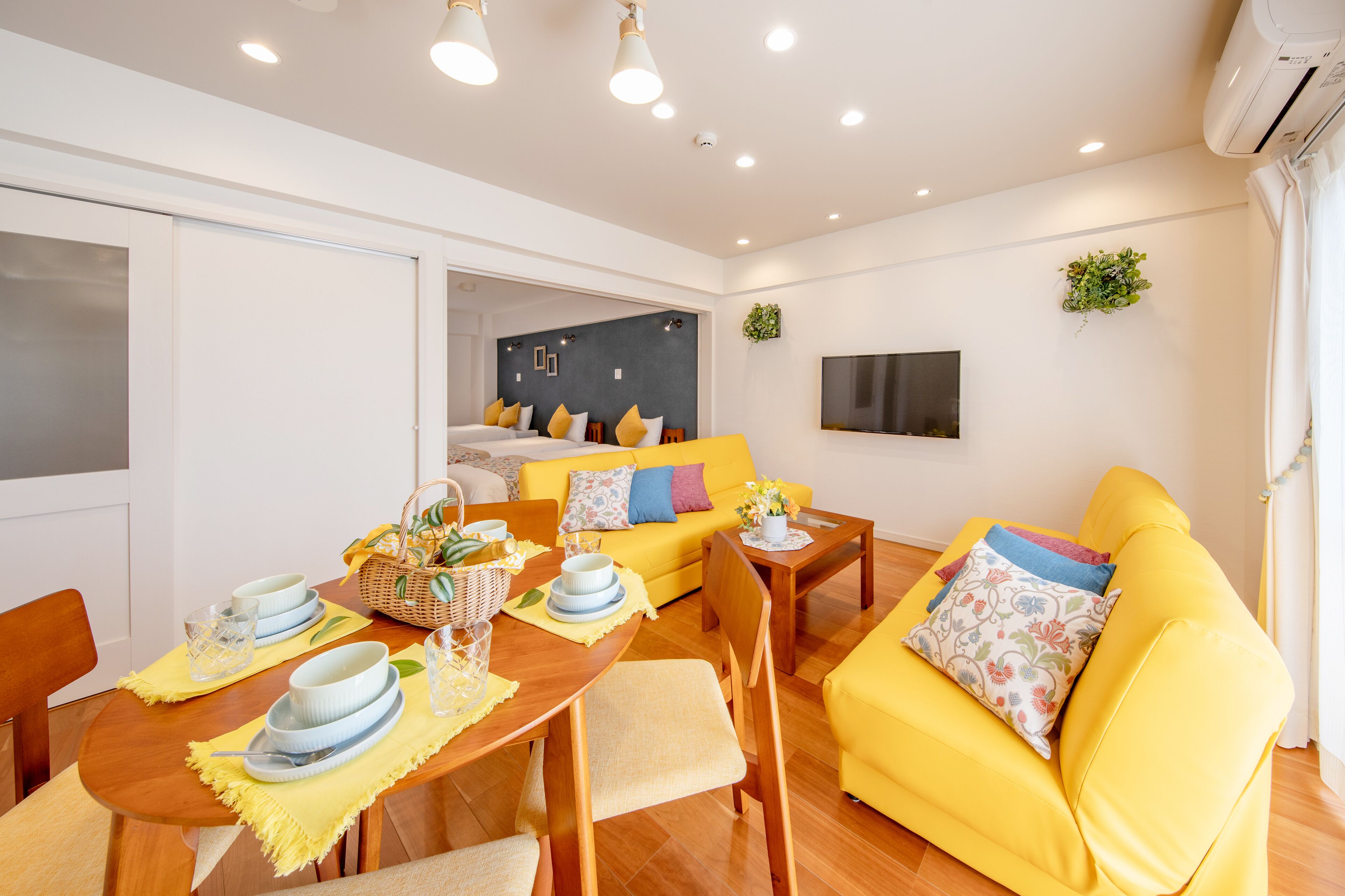 A modern living room with a yellow sofa, a dining table set with plates and glasses, and a TV mounted on the wall.