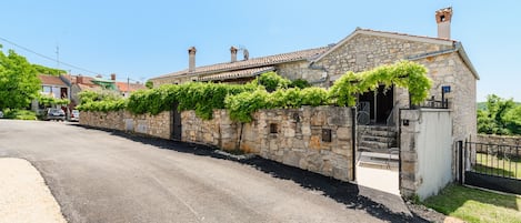 A stone building with a tiled roof, a stone wall with greenery, and a paved road.