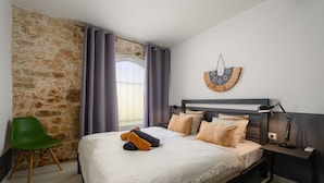 A bedroom with a stone wall, a bed with multiple pillows, a green chair, and a bedside table with a lamp.