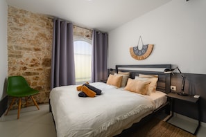 Une chambre avec un mur en pierre, un lit recouvert de plusieurs coussins, un fauteuil vert et une table de chevet avec une lampe.