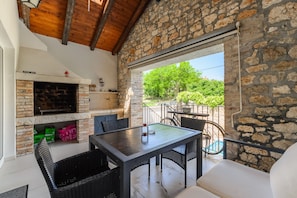 A rustic outdoor seating area with a stone wall, a fireplace, and a view of greenery.