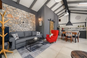 Un salon moderne avec un mur en pierre, un canapé gris, un fauteuil rouge, une table basse et un coin cuisine comprenant une table à manger et des chaises.