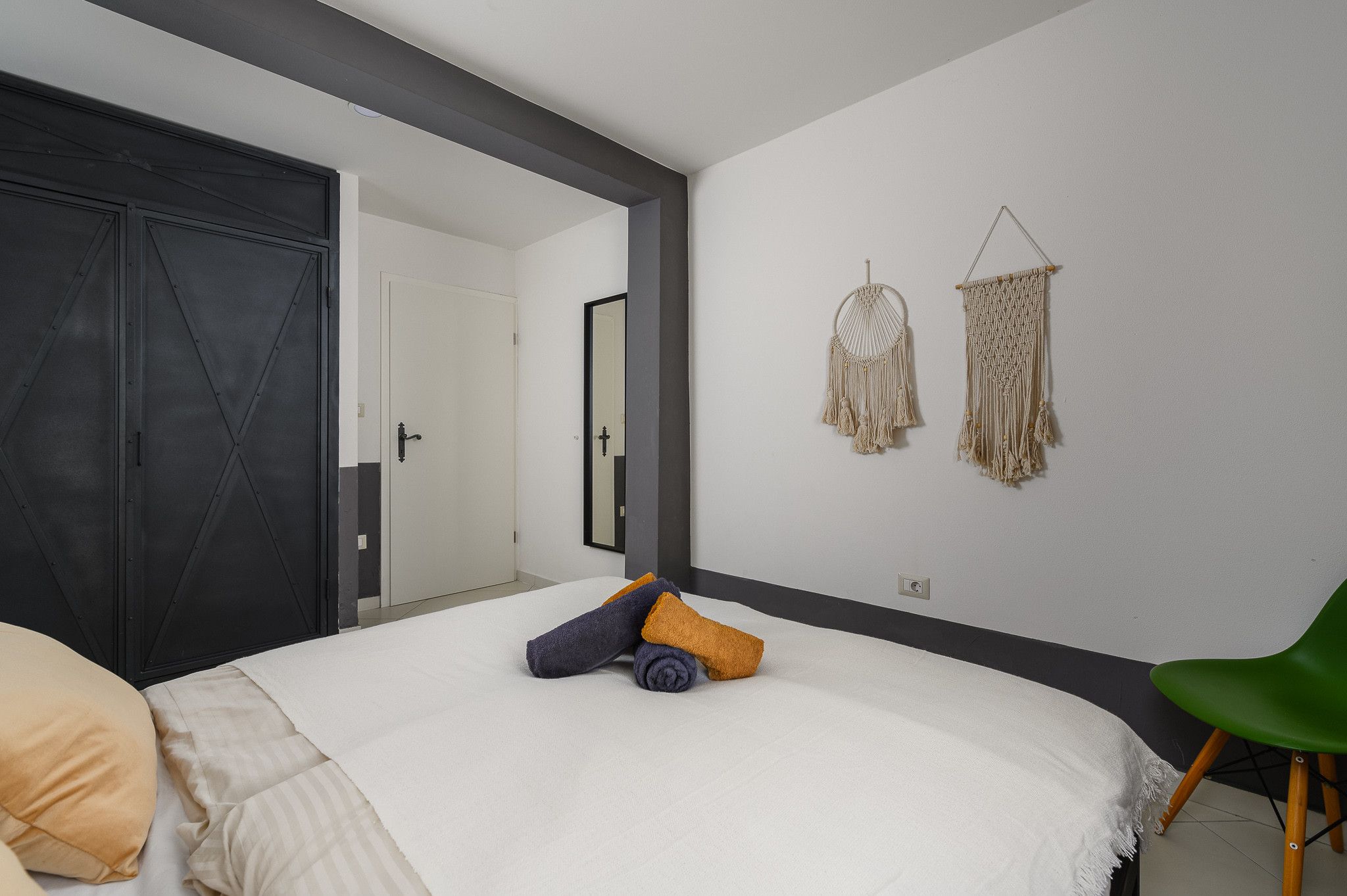 Une chambre à coucher avec un lit, un fauteuil vert, deux décorations murales en macramé et une porte.
