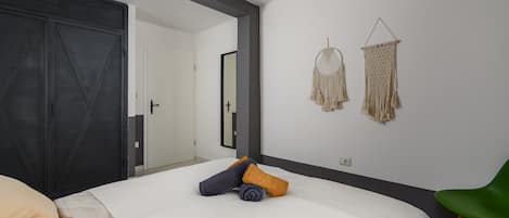 Une chambre à coucher avec un lit, un fauteuil vert, deux décorations murales en macramé et une porte.