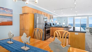 A modern kitchen with wooden cabinets, stainless steel appliances, and a dining area with a blue rug and wooden chairs. - Promo! Second Floor Oceanfront Condo with Pool, Hot Tub, Wifi and Much More! (Depoe Bay)