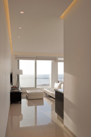 A modern hotel room with a large window offering a sea view, a white sofa, a small table with a lamp, and a flat-screen TV.