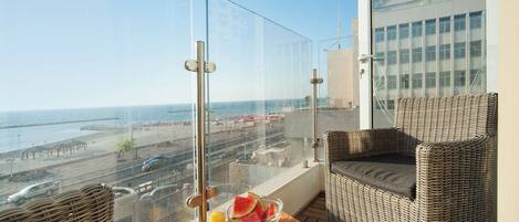 Un balcón con mobiliario de mimbre, una mesa con un frutero y bebidas, y vistas a la playa y al paisaje urbano.