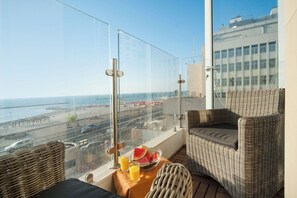 A balcony with wicker furniture, a table with a bowl of fruit and drinks, and a view of the beach and cityscape.