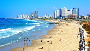 Une plage où des gens se prélassent au soleil et se baignent, un ciel dégagé et, au loin, le profil d’une ville.