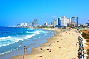 Una spiaggia con persone che prendono il sole e nuotano, cielo sereno e uno skyline cittadino sullo sfondo.