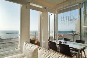 A modern interior with a glass dining table, white leather sofa, and a view of the sea.