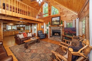 Une cabane en bois spacieuse avec une chambre à coucher en mezzanine, une cheminée en pierre et un grand salon doté d’une autre cheminée et d’un téléviseur.