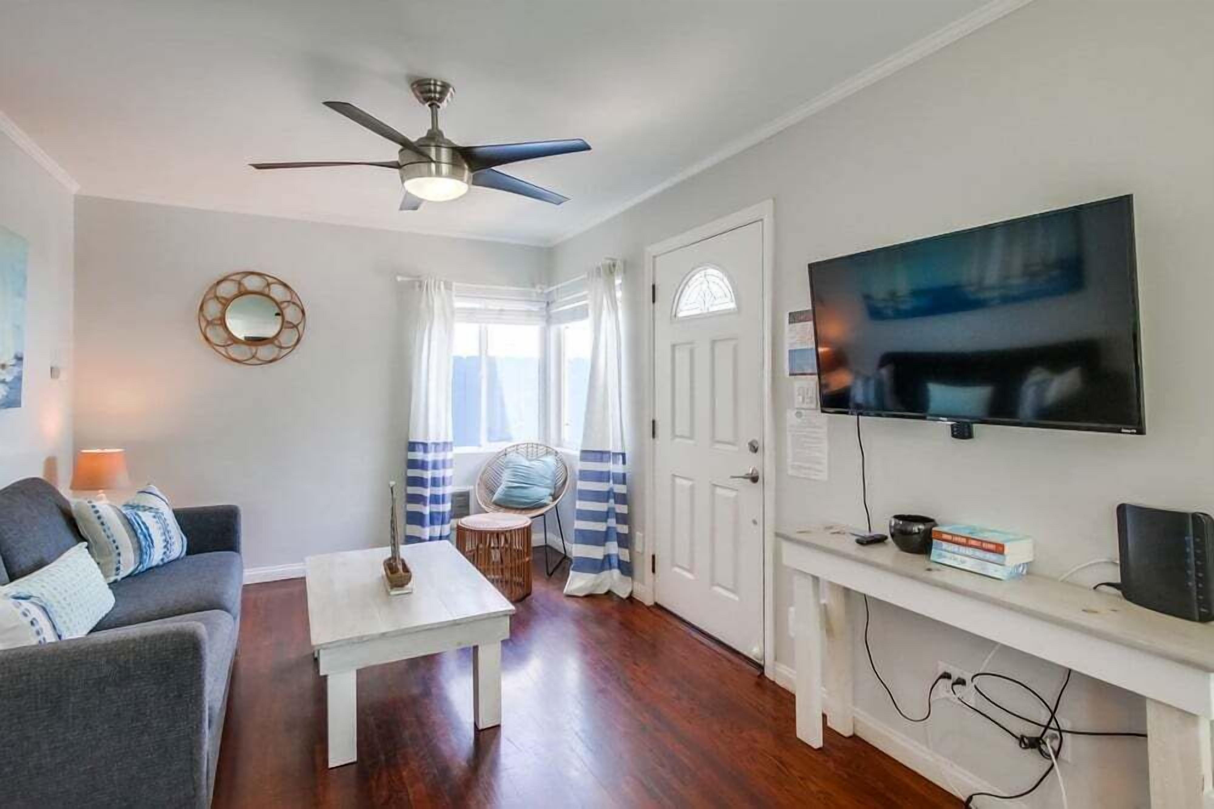 Un salon doté d’un ventilateur de plafond, d’un téléviseur fixé au mur, d’une table basse blanche, d’un canapé avec des coussins et d’une fenêtre avec des rideaux.