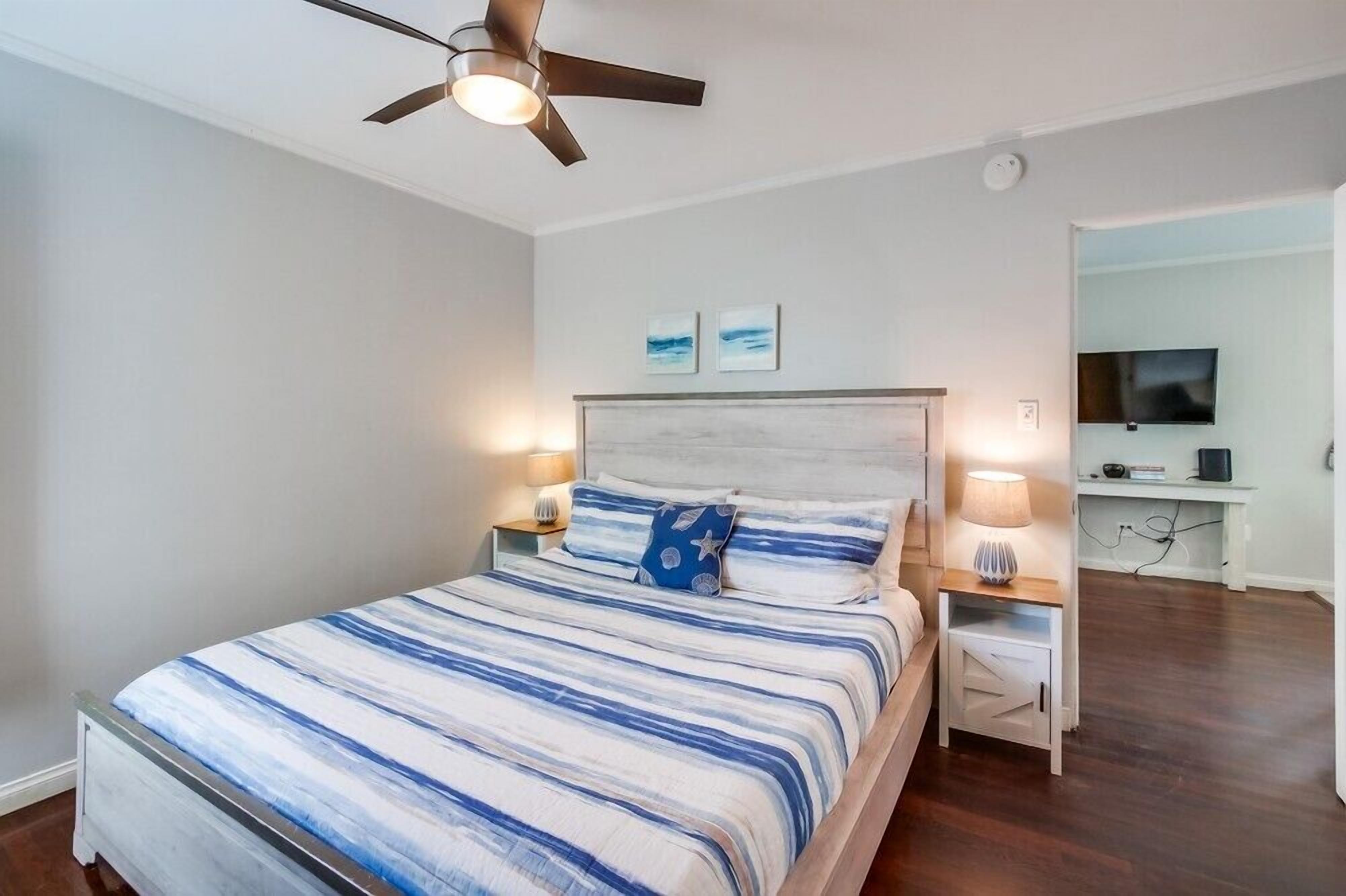 Une chambre à coucher comprenant un lit, des tables de chevet, un ventilateur de plafond, un téléviseur et des œuvres d’art accrochées au mur.