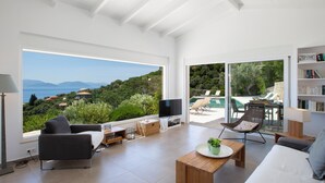 A modern living room with a large window offering a view of the sea and hills. - Villa Koumaria - Secluded Elegance with Amazing Sea Views & Infinity Pool (Sivota)