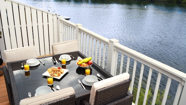 Une table à manger, dressée pour le petit-déjeuner, entourée de chaises en osier, sur une terrasse dotée d’une balustrade blanche, qui offre une vue sur une rivière paisible.