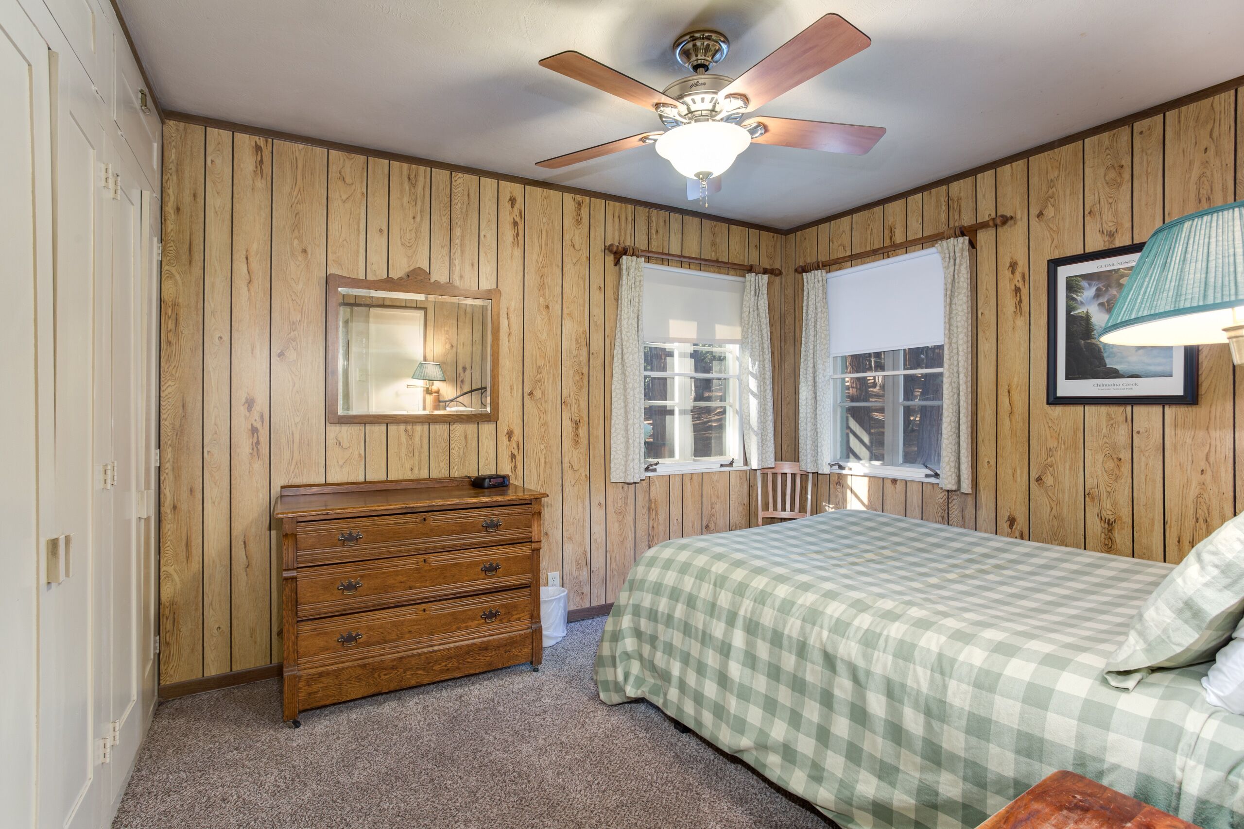Een slaapkamer met houten paneelwanden, een plafondventilator, een bed met een geruit beddegoed, een houten dressoir en een raam met gordijnen.