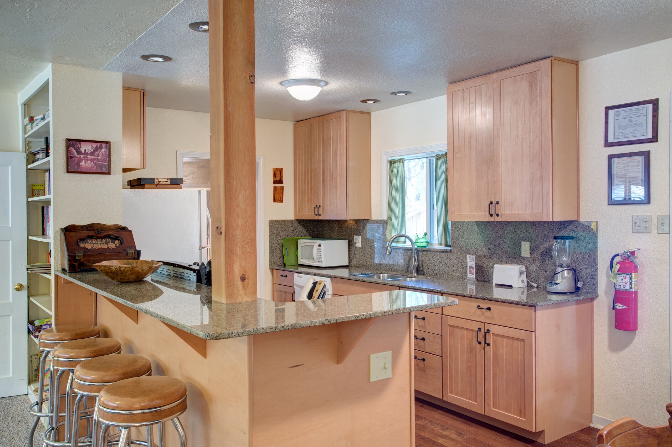 A kitchen with wooden cabinets, a granite countertop, a microwave, a toaster, and a blender. There are bar stools and a window with curtains.