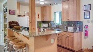 Eine Küche mit Holzschränken, einer Granit-Arbeitsplatte, einer Mikrowelle, einem Toaster und einem Mixer. Es gibt Barhocker und ein Fenster mit Vorhängen.